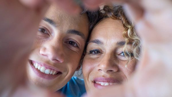 Portrait of couple of mom and son together a home making a heart love shape with their fingers and hands with the faces at the background. Affectionate teenager and woman enjoying