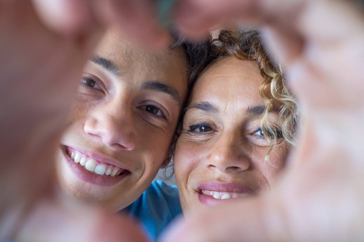 Portrait of couple of mom and son together a home making a heart love shape with their fingers and hands with the faces at the background. Affectionate teenager and woman enjoying